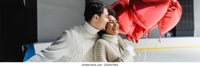 Joyful interracial couple in warm clothes holding red heart shaped balloons on ice rink, banner  - Shutterstock ID 2232927001