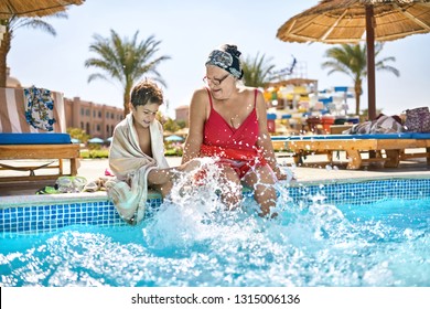 Joyful kid with his grandma are shaking their feet in the water while sitting on the edge of the pool on the sunny background of the hotel. Lady wears a red swimsuit with headband and glasses.