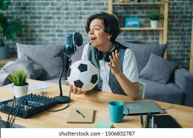 Joyful kid recording audio about football holding ball talking in microphone at home sitting at table alone. Youth lifestyle, sports fans and technology concept. - Powered by Shutterstock