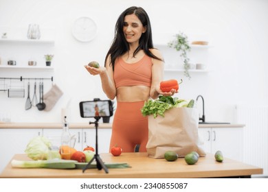 Joyful lady in sportswear holding avocado and sweet pepper while creating online journal using cell phone on tripod. Athletic female blogger explaining benefits of different nutritios vegetables.