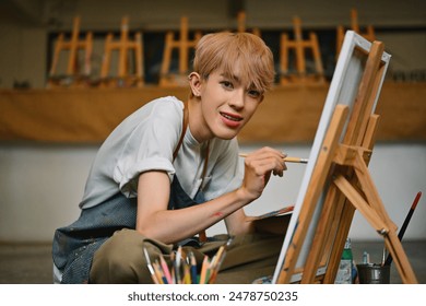 A joyful LGBT artist with blonde hair, wearing a paint-splattered striped apron, paints on a canvas in a bright, spacious art studio filled with light, easels, and various art supplies. - Powered by Shutterstock