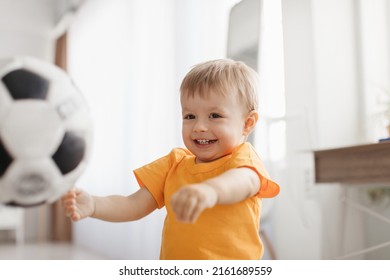 Joyful Little Boy Playing With Football Ball, Having Fun And Enjoying Game At Home, Living Room Interior. Adorable Toddler Kid Throwing The Ball And Smiling