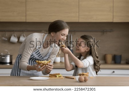 Joyful little kid girl giving mom sandwich to bite. Happy mother and daughter cooking snack for lunch together in kitchen, tasting meal, having fun, smiling, enjoying eating, homemade culinary