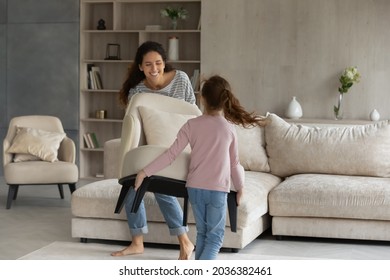 Joyful little kid girl helping smiling hispanic mother moving armchair in modern living room, enjoying renovating stylish apartment. Happy two gen female family homeowners decorating house together.