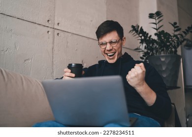 Joyful male entrepreneur in casual clothes and glasses sitting on sofa at home and enjoying good news on netbook with fist up gesture of success - Shutterstock ID 2278234511