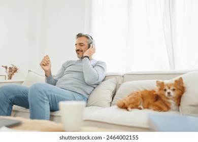 Joyful man with closed eyes feeling the music on headphones, cozying up with his chihuahua dog on a couch at home - Powered by Shutterstock