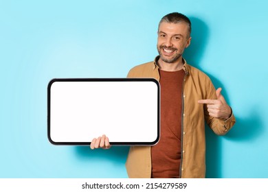 Joyful Man Holding Big Mobile Phone And Pointing Finger At Empty Smartphone Screen Advertising Offer Or App Standing Over Blue Background In Studio. Check This Great Application. Mockup