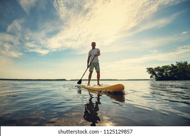 Joyful man is training  SUP board in large river on a sunny morning against a blue sky background . Stand up paddle boarding - awesome active outdoor recreation. Wide angle, backlight.
