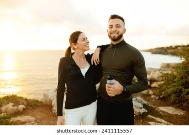 Joyful man and woman in activewear, sharing light moment holding water bottle and embracing against sunset backdrop at seaside - Powered by Shutterstock