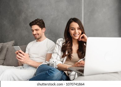 Joyful Man And Woman Sitting Together On Couch In Gray Interior While Using Laptop And Smartphone