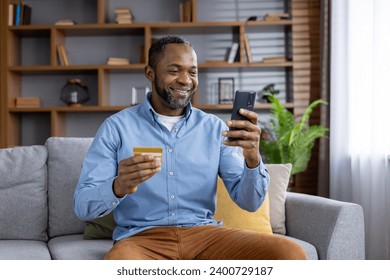 Joyful mature man at home on sofa with phone and bank credit debit card in hands, satisfied african american man in living room chooses gifts in online store, uses app on smartphone, transfers money. - Powered by Shutterstock