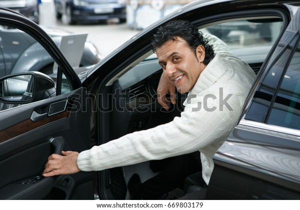 Joyful mature man smiling to the camera over his\
shoulder closing car door while sitting in an automobile male\
buying a car choosing comfort business sales offer driving journey\
travel vehicle