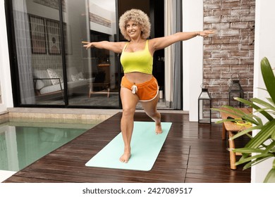 Joyful mature woman with curly hair doing yoga outdoors near a swimming pool, promoting health and wellbeing. - Powered by Shutterstock