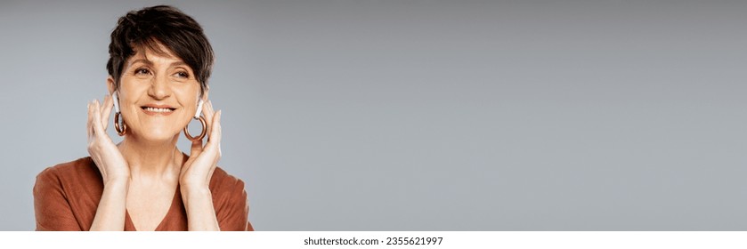 joyful mature woman in earphones listening music on grey, brown jacket, fall fashion,  banner - Shutterstock ID 2355621997