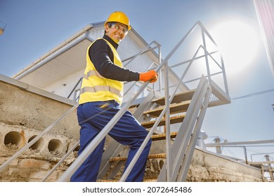 Joyful matured man in safety helmet looking at camera and smiling while walking up staircase at industrial plant - Powered by Shutterstock