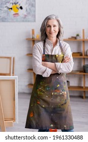 joyful middle aged artist in apron standing with crossed arms near easel