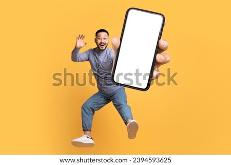 Joyful middle aged Asian man holding large cellphone with blank screen, leaping mid air in studio, humorous and dynamic, perfect for technology and app mockups, yellow background