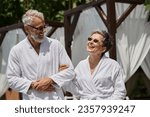 joyful middle aged couple in sunglasses and robes walking in luxury resort, wellness retreat concept