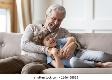 Joyful middle aged hoary man playing having fun with happy small grandchild in living room. Excited older senior grandfather tickling laughing kid boy, relaxing together on comfortable sofa. - Powered by Shutterstock