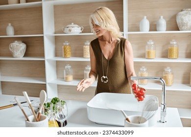 joyful middle aged woman washing cherry tomatoes and looking at fresh greenery in modern kitchen - Powered by Shutterstock