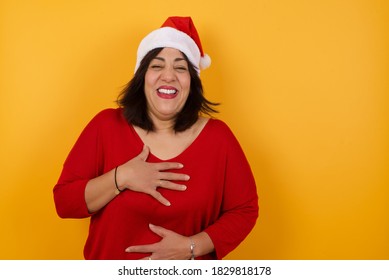 Joyful Middle Aged Woman Wearing Christmas Hat  Keeps Hands Crossed, Laughs At Good Joke, Wears Red Sweater, Standing Indoors. 