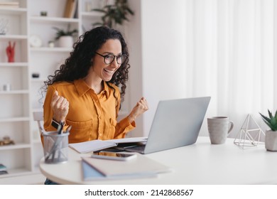 Joyful Middle Eastern Woman Shaking Hands Using Laptop Computer Celebrating Business Success Working Online Sitting At Desk At Home. Remote Job And Freelance, Big Luck Concept. Side View