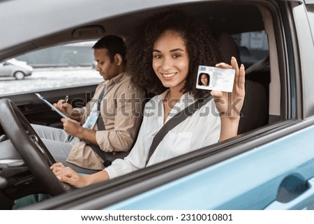 Joyful Middle Eastern Woman Showcasing Her Driving License To Camera Through Car Window Sitting Inside, While Professional Instructor Taking Notes After Successful Test Drive. Selective Focus