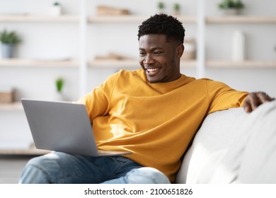 Joyful Millennial Black Guy In Casual Sitting On Couch With Laptop, Surfing On Internet, Watching Movie Or Playing Game Online, Looking At Notebook Screen And Smiling, Home Interior, Copy Space