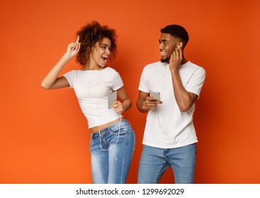 Joyful Millennial Black Man And Woman In Earphones Listening To Music On Cellphones And Smiling To Each Other, Orange Background