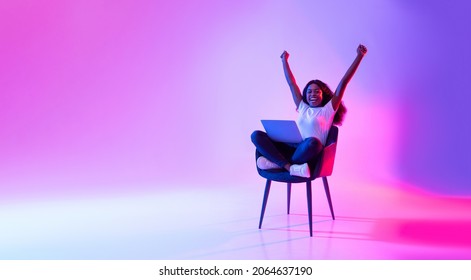 Joyful Millennial Black Woman Sitting In Armchair With Laptop, Celebrating Huge Online Win Or Success In Neon Light, Banner With Copy Space. Young African American Lady Enjoying Big Sale In Web Store