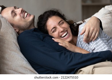 Joyful Millennial Couple Having Fun And Enjoying Closeness At Home, Hugging On Couch, Relaxing, Talking, Laughing, Showing Perfect Teeth. Happy Husband And Wife Resting On Soft Comfortable Couch