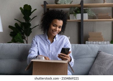 Joyful Millennial Female African Ethnicity Consumer Woman Reading Message In Cellphone Application, Unpacking Carton Box, Leaving Review Satisfied With Positive Online Shopping Experience.