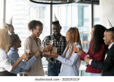 Joyful mixed race african workers in paper party hats congratulating sincere laughing biracial colleague with happy birthday, presenting wrapped gift box, wishing all the best in modern workplace.