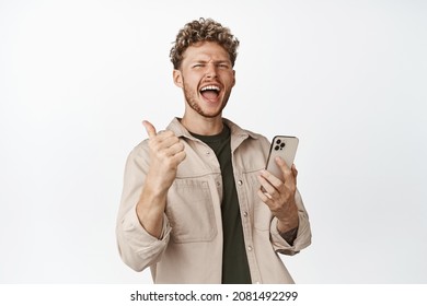 Joyful modern blond guy triumphing with mobile phone in hand, screaming happy and showing thumbs up, celebrating achievement on smartphone, white background