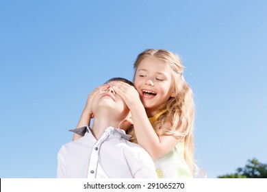 Joyful moment. Pretty little girl holding her hands on the face of boy and closing his eyes while sitting on blanket alfresco