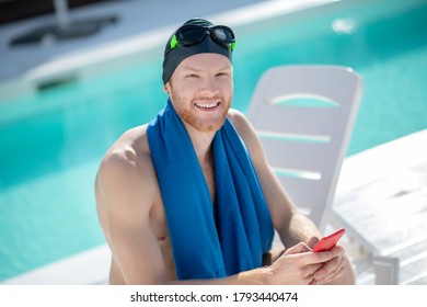 Joyful mood. Smiling young bearded man in black swimming cap and blue towel with smartphone sitting near the pool - Powered by Shutterstock