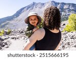 A joyful mother carries her smiling baby boy in a sun hat, enjoying the breathtaking mountain landscape of Crowsnest Pass, Alberta, Canada on a sunny day.