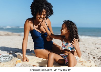 A joyful mother and daughter applying sunscreen while enjoying a sunny day at the beach, emphasizing family time and sun protection. - Powered by Shutterstock