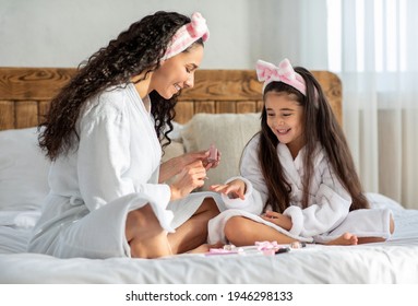 Joyful Mother And Daughter Making Manicure At Home, Wearing Bathrobes, Sitting On Bed And Applying Nail Polish On Hand Nails, Cute Mom And Little Girl Having Beauty Day, Bedroom Interior