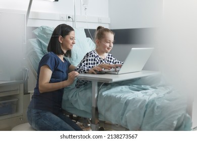 Joyful Mother Enjoying Leisure Time With Smiling Cheerful Daughter While In Pediatric Clinic Ward Room Using Laptop. Mother And Daughter Sitting In Pediatric Hospital Room While Using Computer.