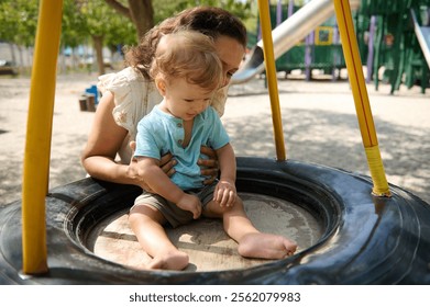 A joyful mother gently holds her child as they play on a tire swing in a vibrant park. - Powered by Shutterstock