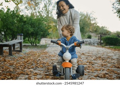 A joyful mother pushes her toddler on a tricycle along a leaf-strewn path in a park. The scene captures the warmth of autumn, laughter, and bonding while enjoying the outdoors together. - Powered by Shutterstock