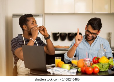 Joyful Multi Ethnic Students Cooking And Having Fun In Kitchen. Young Indian Man And His African American Friend Sitting At Table, Drinking Fresh Juice, Watching Video Recipe, Culinary Show On Laptop.