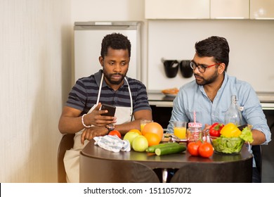 Joyful Multi Ethnic Students Cooking And Having Fun In Kitchen. Young Indian Man And His African American Friend Sitting At Table, Drinking Fresh Juice, Watching Video Recipe, Culinary Show On Laptop.