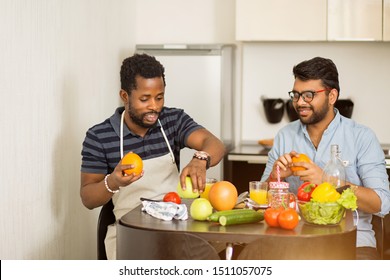 Joyful Multi Ethnic Students Preparing Healthy Food And Having Fun In Kitchen. African American Guy And His Indian Friend Sitting At Table Watching Cooking Show And Laughing. Cooking And Technology.