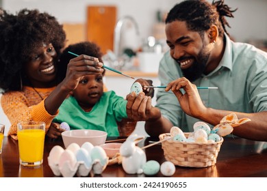 A joyful multicultural family is sitting at the table at home together and painting easter eggs on easter saturday. Young dad is holding an egg while mother is teaching their son how to color eggs. - Powered by Shutterstock