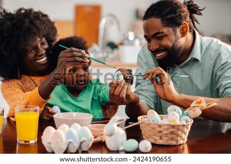 A joyful multicultural family is sitting at the table at home together and painting easter eggs on easter saturday. Young dad is holding an egg while mother is teaching their son how to color eggs.