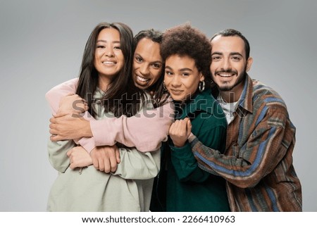 joyful multicultural friends in trendy outfit embracing and looking at camera isolated on grey