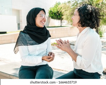 Joyful Multiethnic Friends And Colleagues Discussing Good News Outside. Women In Office Suits And Hijab Sitting Outdoors, Chatting And Laughing. Friendly Colleagues Concept