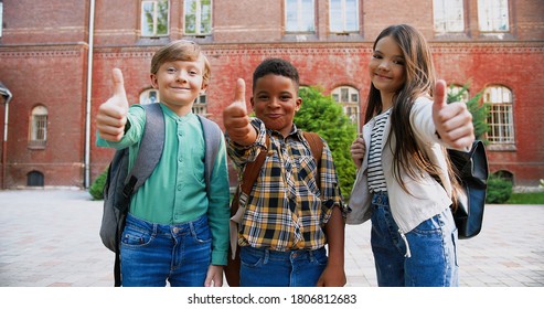 Joyful multi-ethnic junior students with school bags standing in front of school and giving thumbs up. Pretty Caucasian girl smiling outdoor. African American cute boy in good mood. Portrait concept